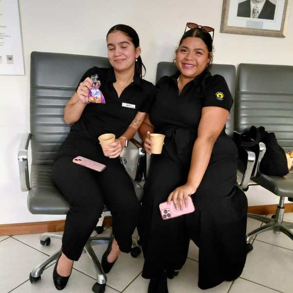 Two women sitting in chairs, smiling, holding cups, and a framed photo on the wall.