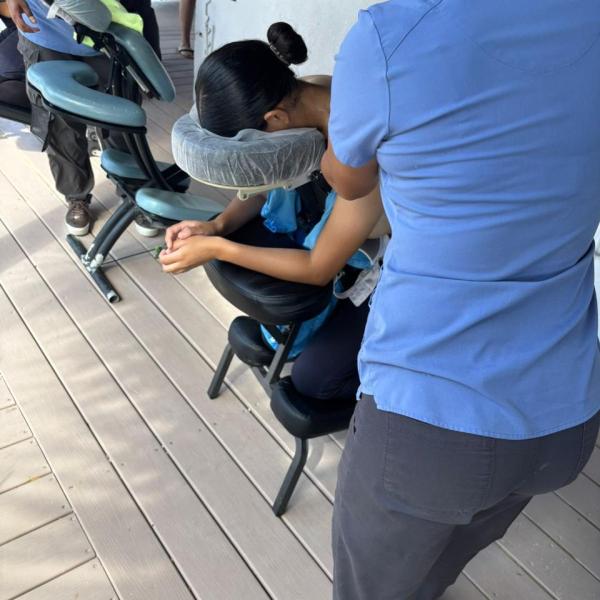 Person in blue shirt gives seated massage to someone on a massage chair on a wooden deck.