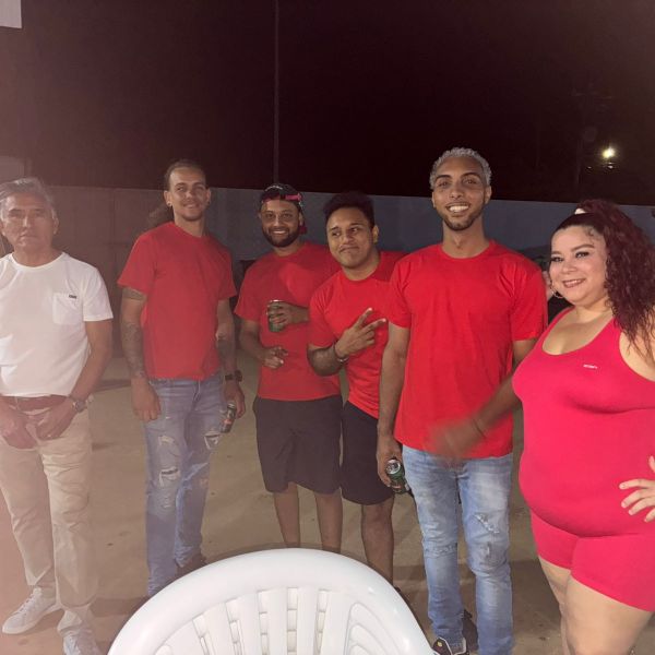 Group of people posing at a gathering, wearing mostly red and white outfits, with plastic chairs in the foreground.