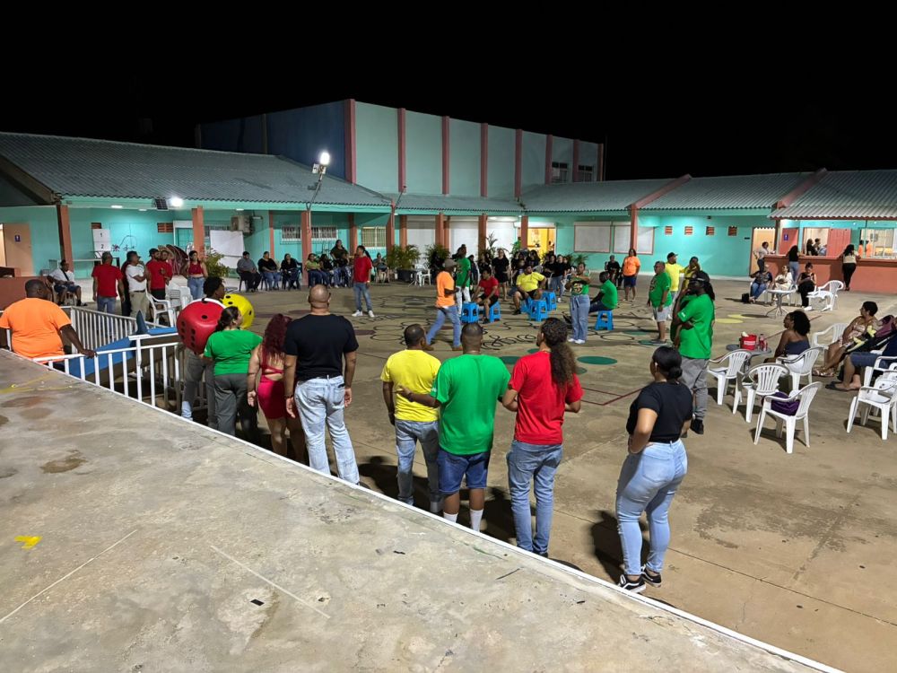 People in colorful shirts gathered outdoors for an event at night, with chairs and buildings in the background.