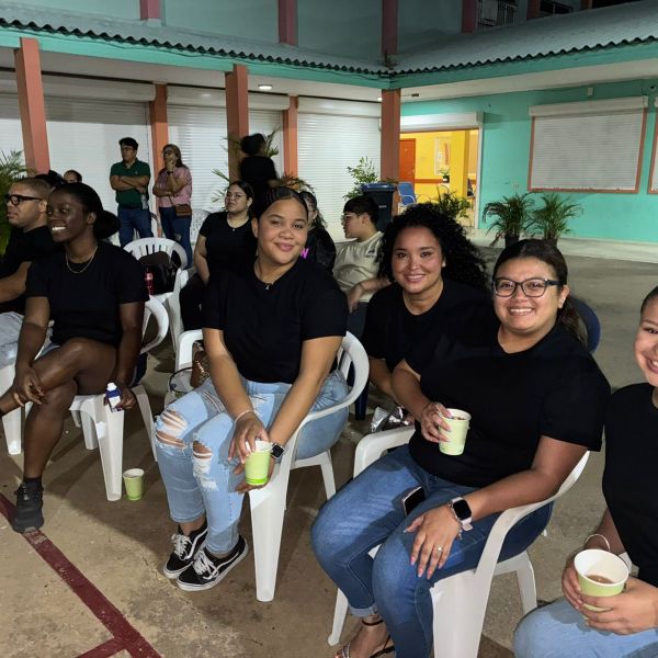 Group of people sitting in outdoor courtyard on plastic chairs, wearing black shirts, smiling and holding cups.