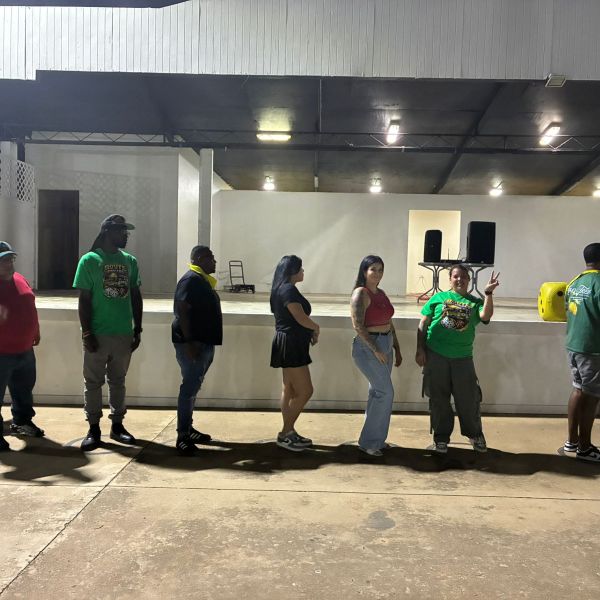 A diverse group of people stands in line indoors on a concrete floor, with a stage in the background.