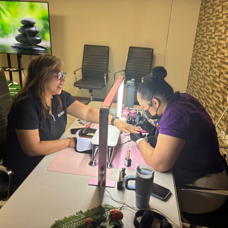 Two women in a salon, one giving a manicure at a table with nail tools.
