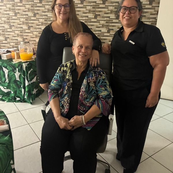 Three women smiling, one seated, against a brick-patterned wall and green tablecloth with food in the background.