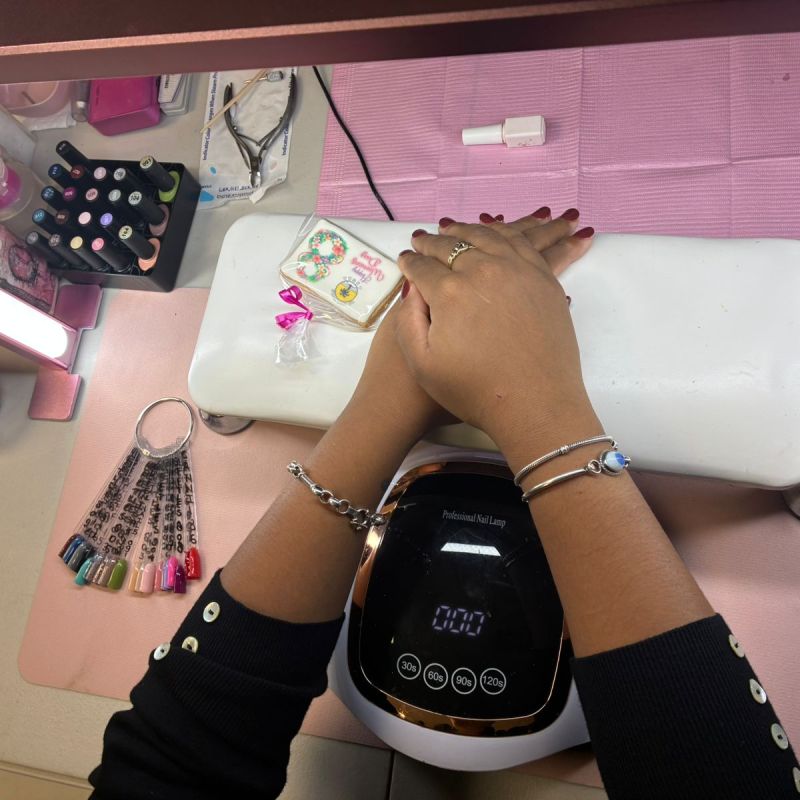 Hands under UV nail lamp with nail polish samples and art tools on a pink mat.