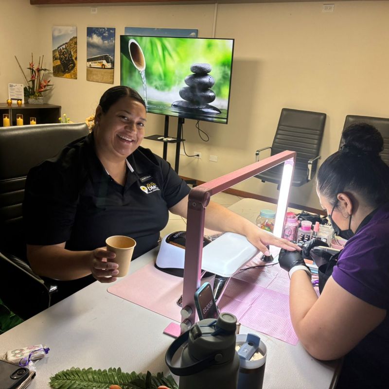 Two women at a table, one getting her nails done, both smiling. Decor includes a zen rock picture.