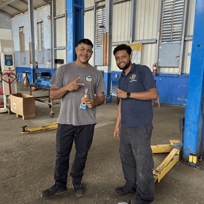 Two men standing in a workshop, smiling and pointing at the camera.