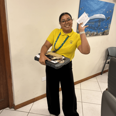 Person in yellow shirt holding a box and paper, standing in a room with a submarine painting.