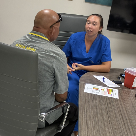 Woman in blue scrubs talks to man in grey shirt at a desk with papers.