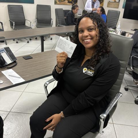 Smiling woman holding a card, sitting in an office with others around.