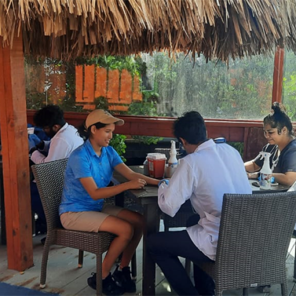 People sitting at tables under a thatched roof, with a waiter serving drinks.