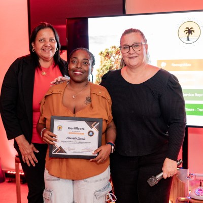 Three women pose with a certificate in front of a screen during an indoor event.