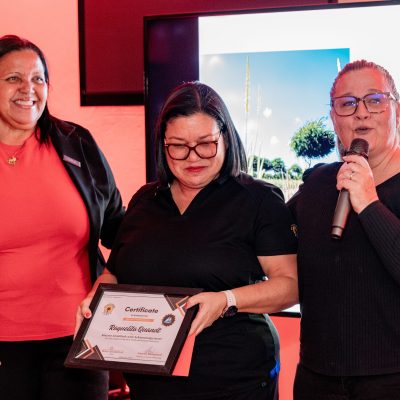Three women, one holding a certificate, stand in front of a screen showing 'De Palm Tours Aruba' logo.