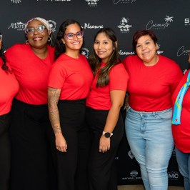 Six women in red shirts and dark pants pose together in front of a branded backdrop.