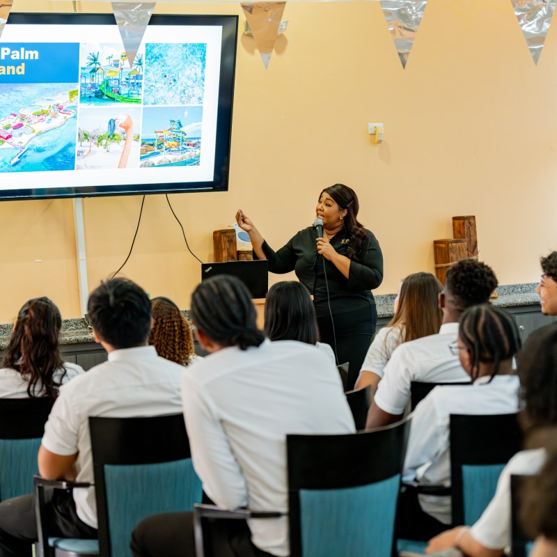 Woman presenting slides about a tropical island to a seated audience in a classroom.