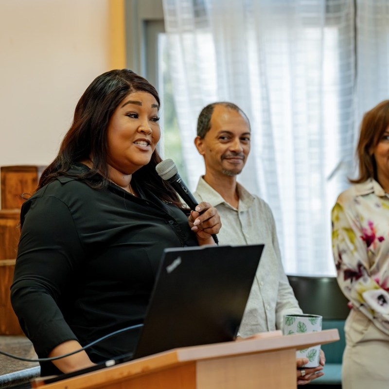 Woman speaking at a podium with microphone, two people standing nearby.