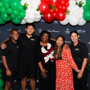Five people smiling under balloon arch with logos on backdrop.