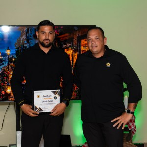 Two men in black shirts, one holding a certificate, standing indoors with a screen behind them.