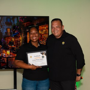 Two people smiling, one holding a certificate, standing indoors beside a large screen displaying a dining scene.