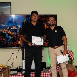 Two men standing indoors; one holds a certificate, another has a microphone.