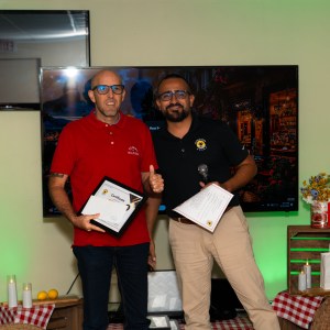 Two men smiling with certificates, standing in a decorated room with TV screens.