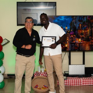 Two men posing with a certificate in a decorated room with balloons and a colorful painting.
