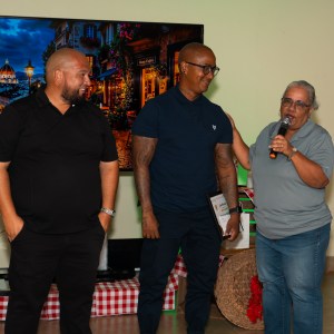 Three people smiling, one holding a microphone, indoors with a large photo in the background.
