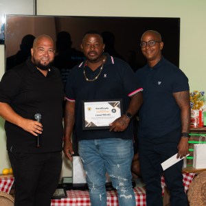 Three men standing, one holding a certificate in front of a TV screen.