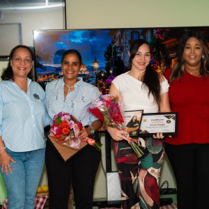 Four women standing, two holding awards and bouquets, smiling indoors.