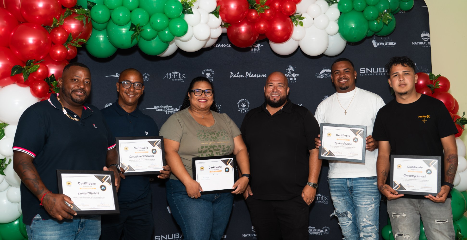 Six people holding certificates, standing under red, green, and white balloon decoration.