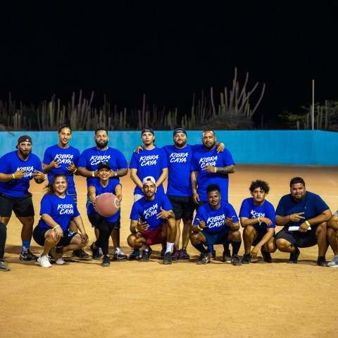 Group photo of a kickball team in blue shirts posing on a field at night.
