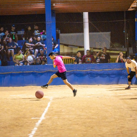 Person in pink shirt kicking a ball on indoor court with spectators watching.