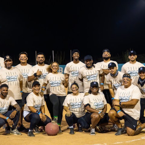 Group of people posing in a softball field at night, wearing matching shirts and casual attire.