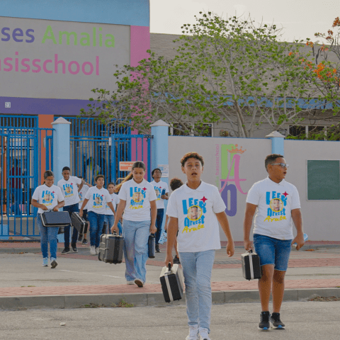 Group of students in matching shirts walking out of a school building carrying cases.