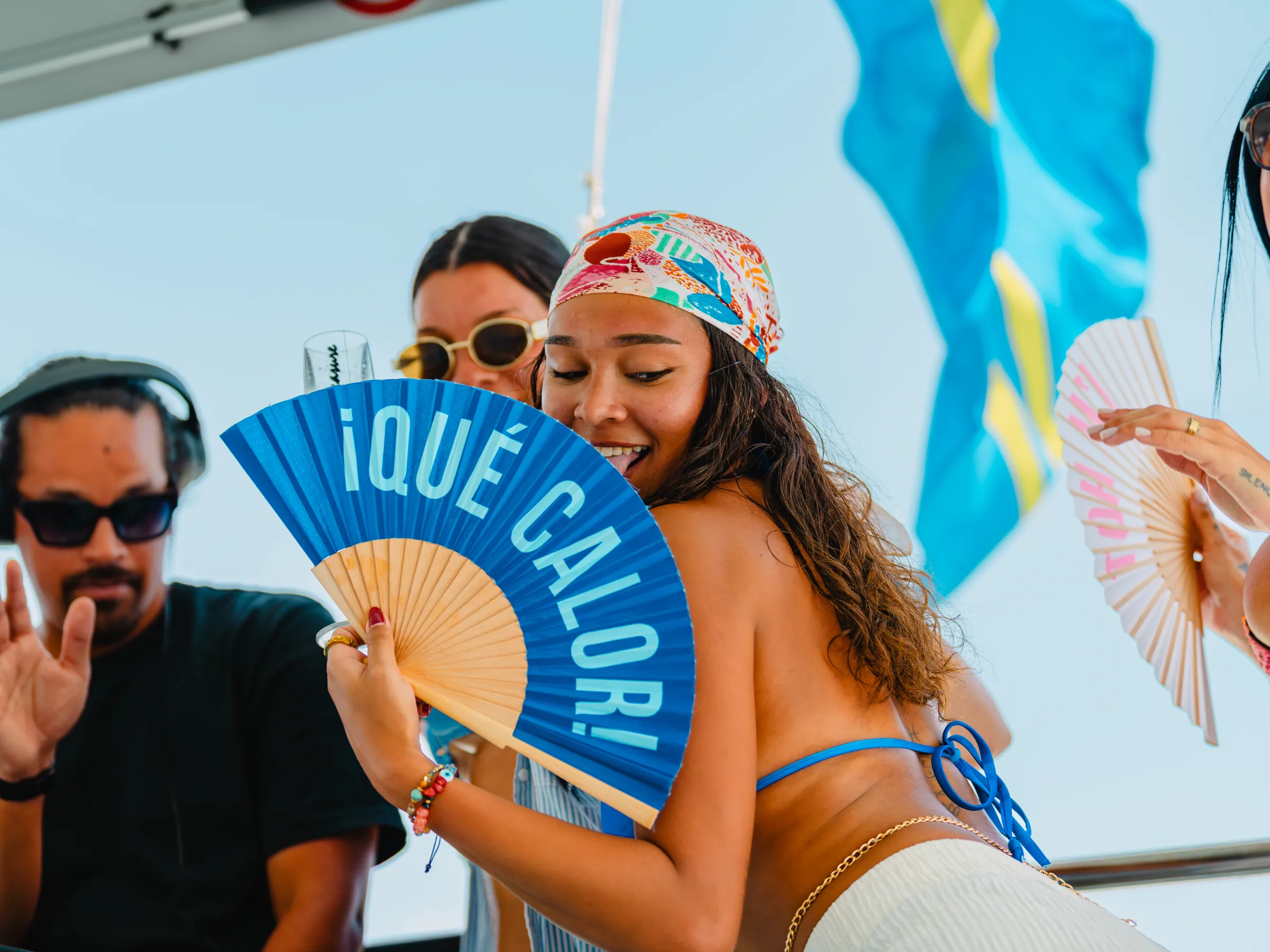 People dancing and enjoying a party on a boat; woman holds a fan with '¡Qué Calor!' text.