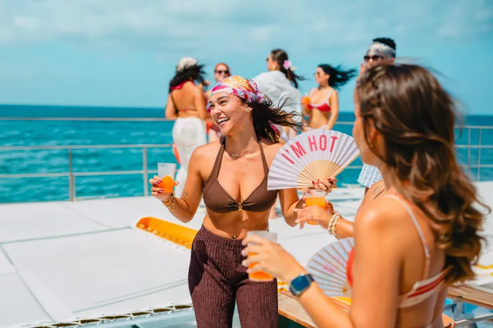 People on a boat enjoying drinks, women wearing swimsuits, clear sky and ocean in the background.