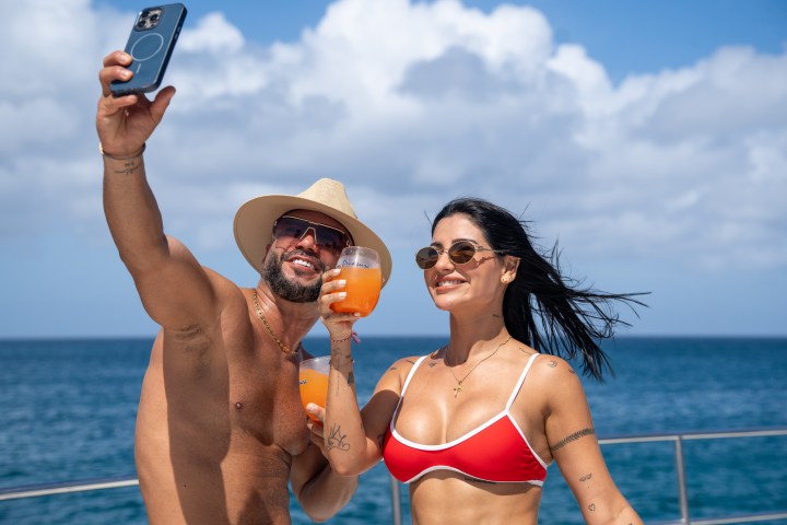Two people on a boat taking a selfie with drinks, ocean in background.