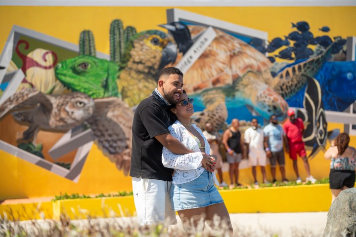 Couple embraces in front of colorful animal mural; people in background.