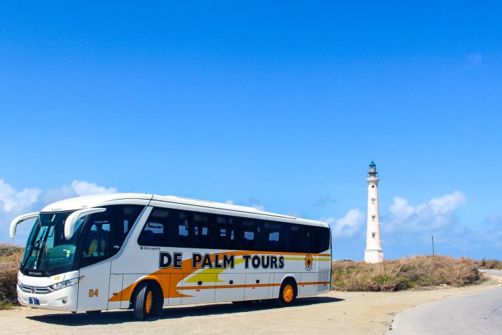 Tour bus parked on a dirt road near a lighthouse under a clear blue sky.
