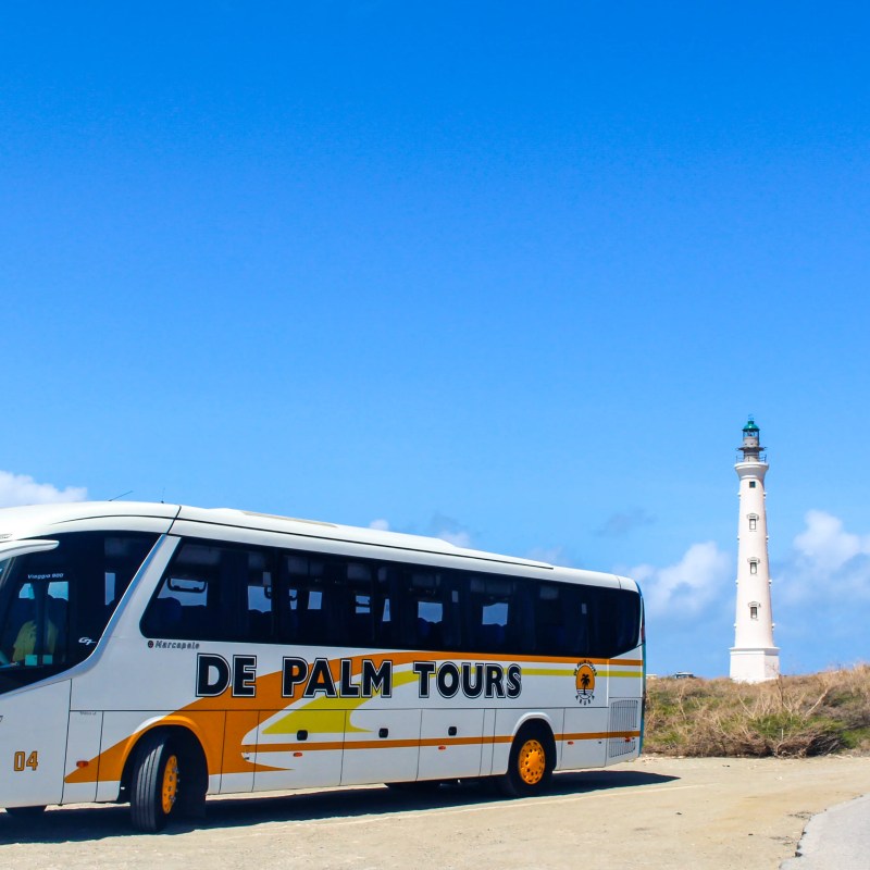 Tour bus parked on a dirt road near a lighthouse under a clear blue sky.