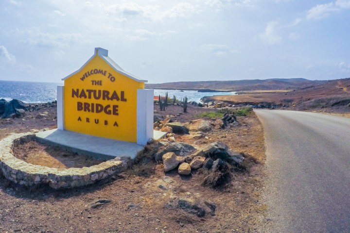 Sign for Natural Bridge Aruba by a coastal road, rocky terrain in the background.