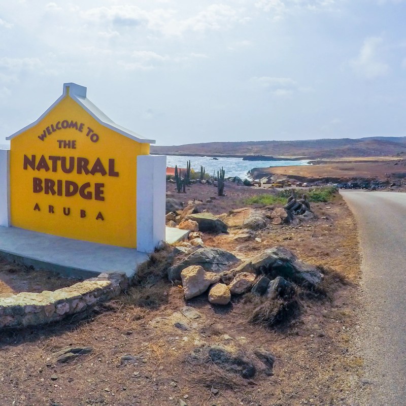 Sign for Natural Bridge Aruba by a coastal road, rocky terrain in the background.