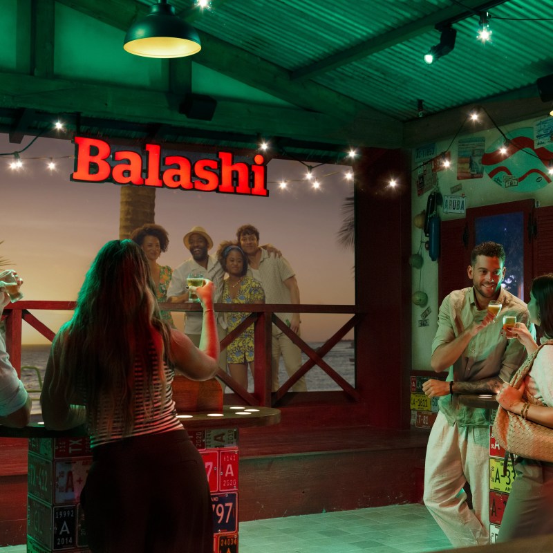 People socializing at a bar under string lights with a Balashi sign.